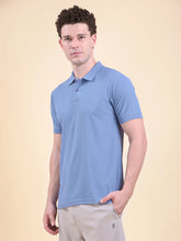 Man wearing a blue polo t-shirt  (DCYPHR Men's Element Polo - Denim Blue) against a beige background facing towards camera posing side ways.